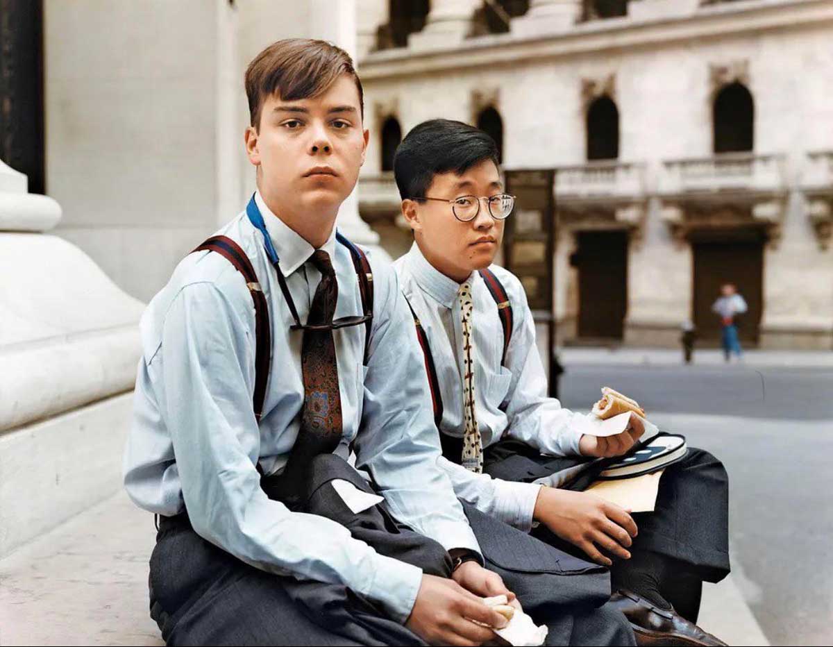 Two young men in dress shirts, ties, and suspenders sit on outdoor steps, eating sandwiches. An old stone building is visible in the background, and both appear to be taking a break.