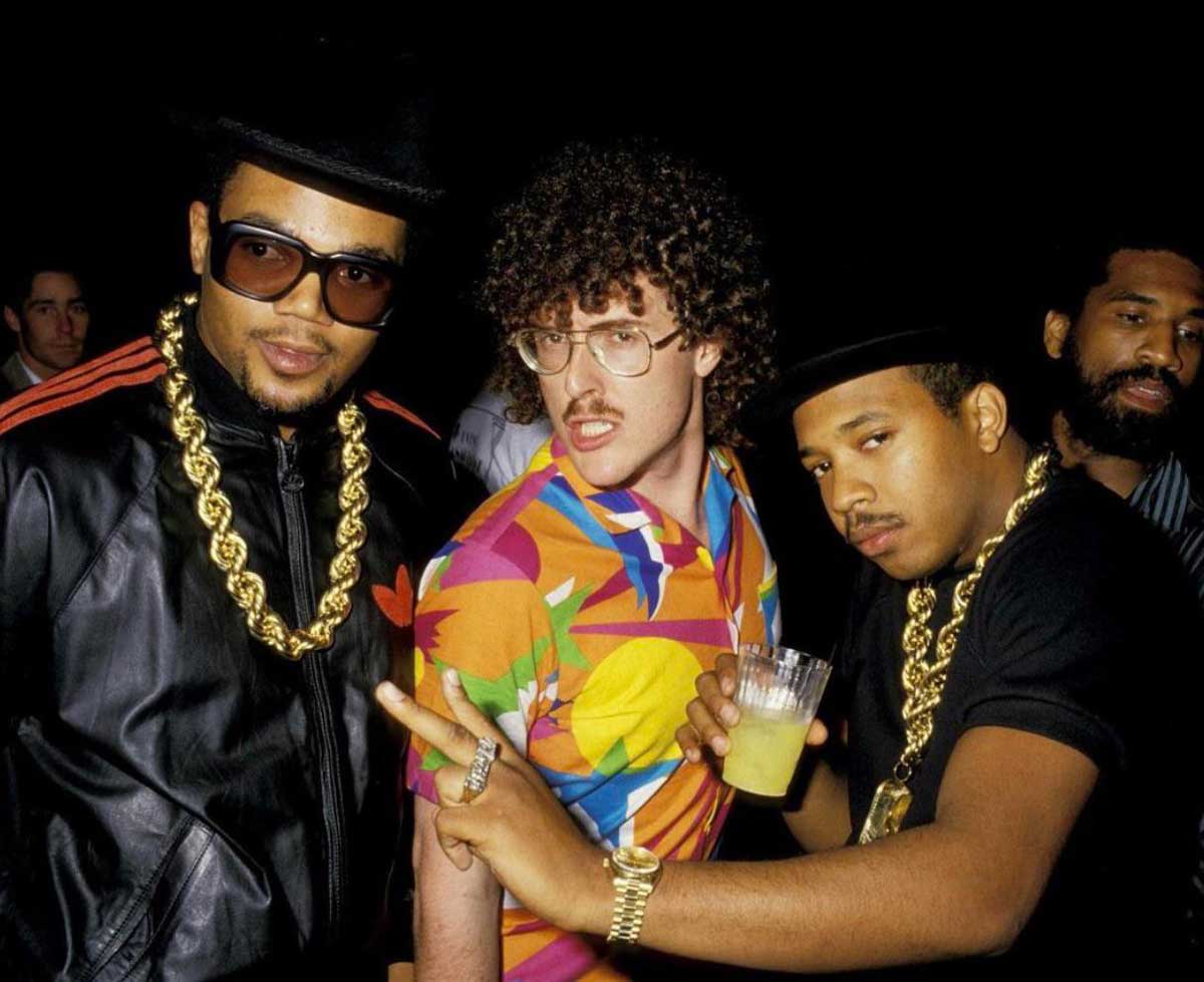 Run DMC and Weird Al Yankovic pose together at a party. Two wear black hats, sunglasses, gold chains, and black outfits, while the third wears glasses and a colorful shirt. One man holds a drink and flashes a peace sign. The background is dark.