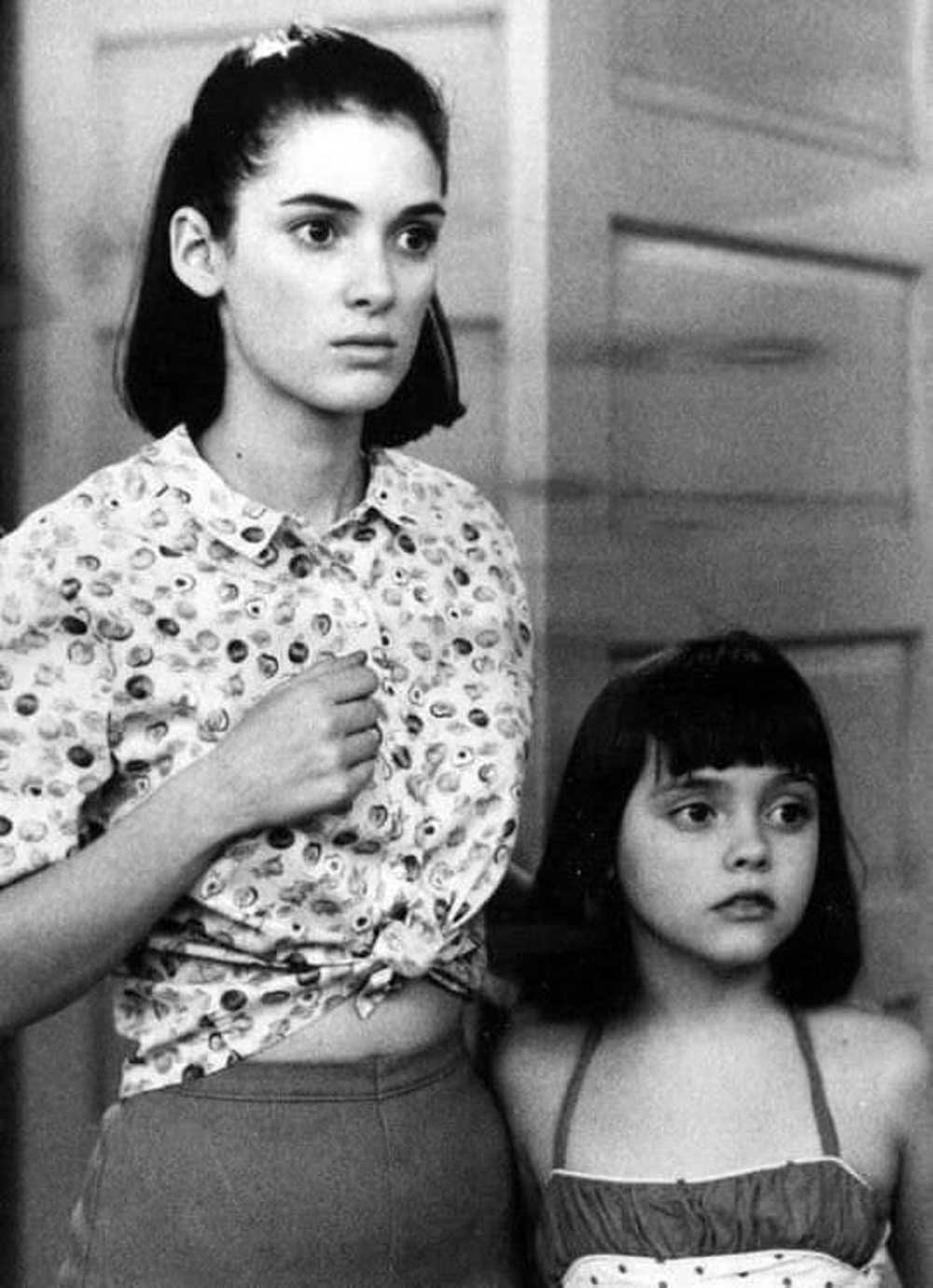 Two girls with dark hair stand together indoors, looking slightly off to the side with serious expressions. The older girl wears a patterned shirt, and the younger wears a sleeveless top. The image is in black and white.
