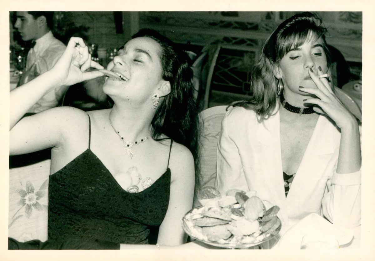 Two young women sit at a table, both smoking cigarettes. One woman wears a black dress and smiles with her eyes closed, while the other wears a white jacket and looks relaxed. A plate of cookies is in front of them.