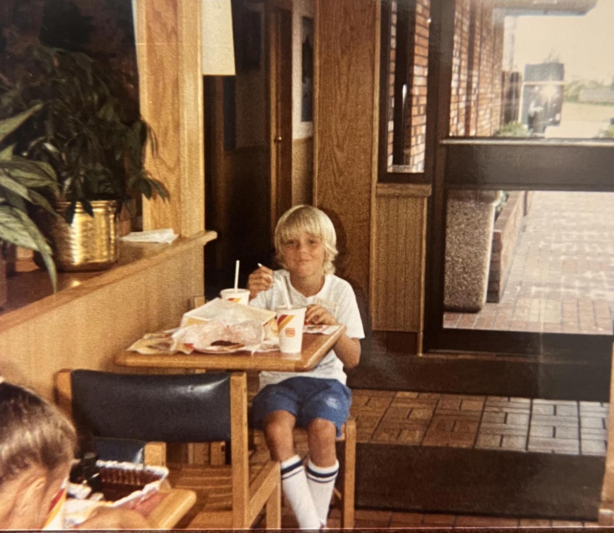 A young boy with blonde hair sits alone at a table in a fast food restaurant, eating a meal with drinks and food wrappers in front of him. He wears blue shorts, a white shirt, and knee-high socks with stripes.