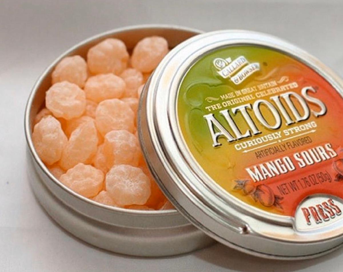 An open tin of Altoids Mango Sours shows pale orange, round candies inside. The lid, partially open, displays the Altoids logo and Mango Sours flavor in bold lettering with vibrant colors.