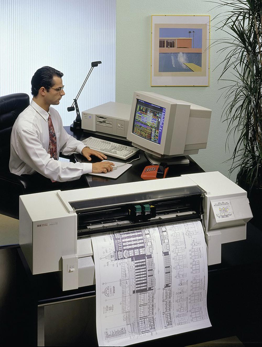 A man in an office sits at a desk using a vintage computer and keyboard. A large HP plotter printer is printing architectural blueprints. A framed artwork hangs on the wall behind him.