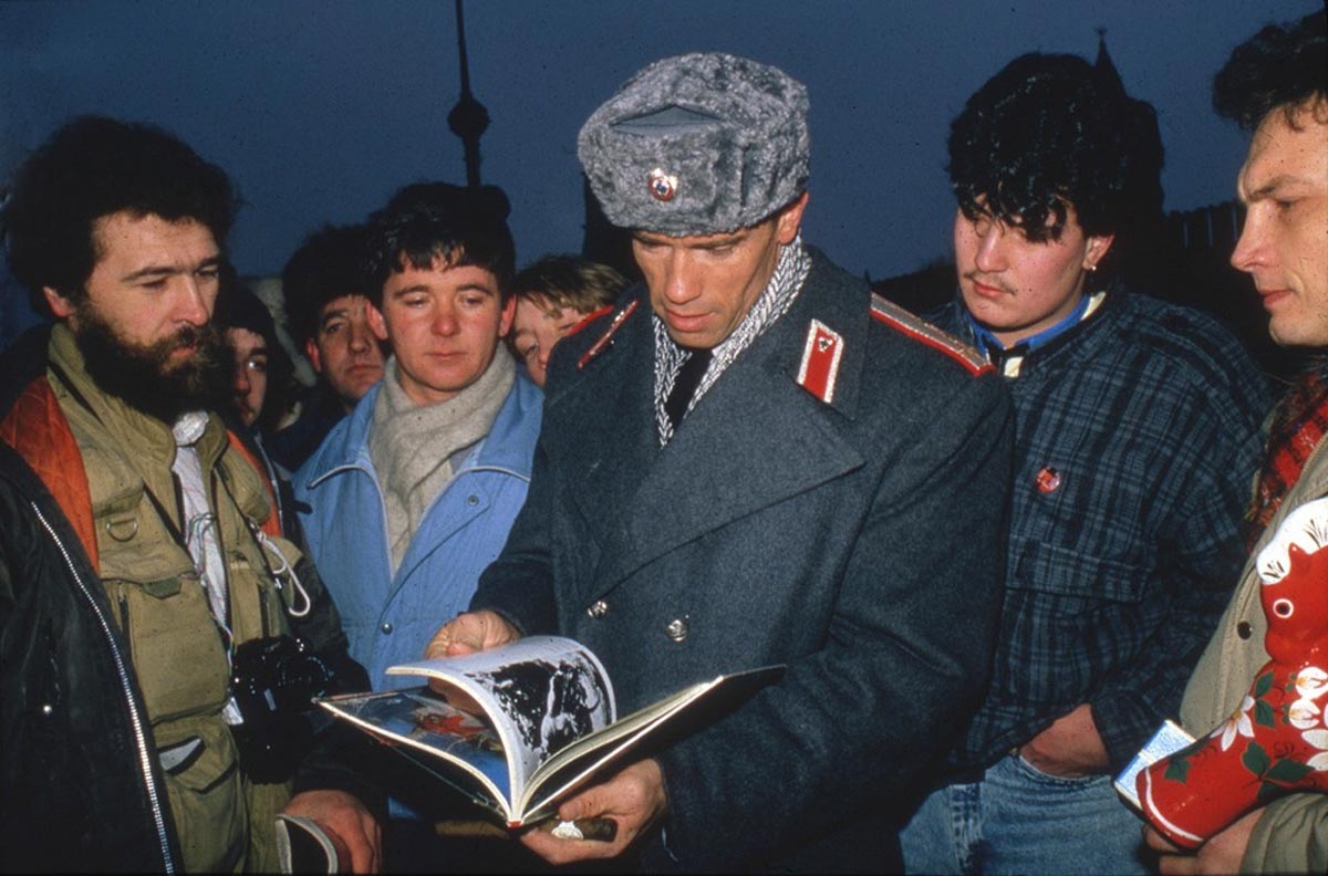 A group of men gather closely in winter clothing, watching as a man in a gray Soviet military-style coat and hat shows them pages from a large open photo book. It appears to be evening or dusk.