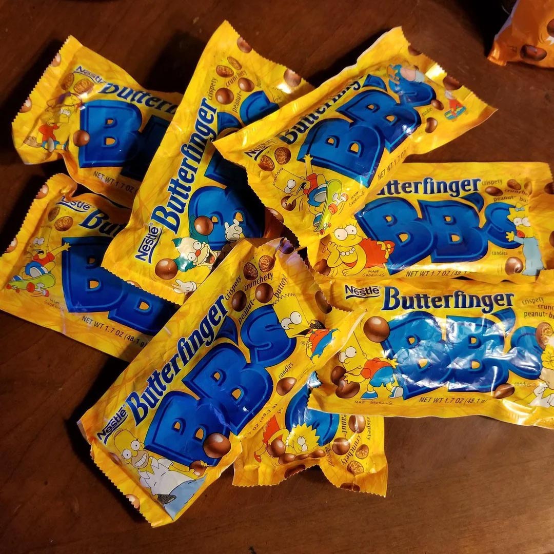 A pile of yellow Butterfinger BBs candy bags featuring cartoon characters from The Simpsons, displayed on a wooden surface.