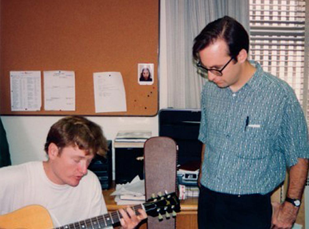 Two men are in an office. One is sitting and playing an acoustic guitar while the other, wearing glasses and a striped shirt, stands nearby watching. There’s a corkboard with papers and a photo on the wall behind them.