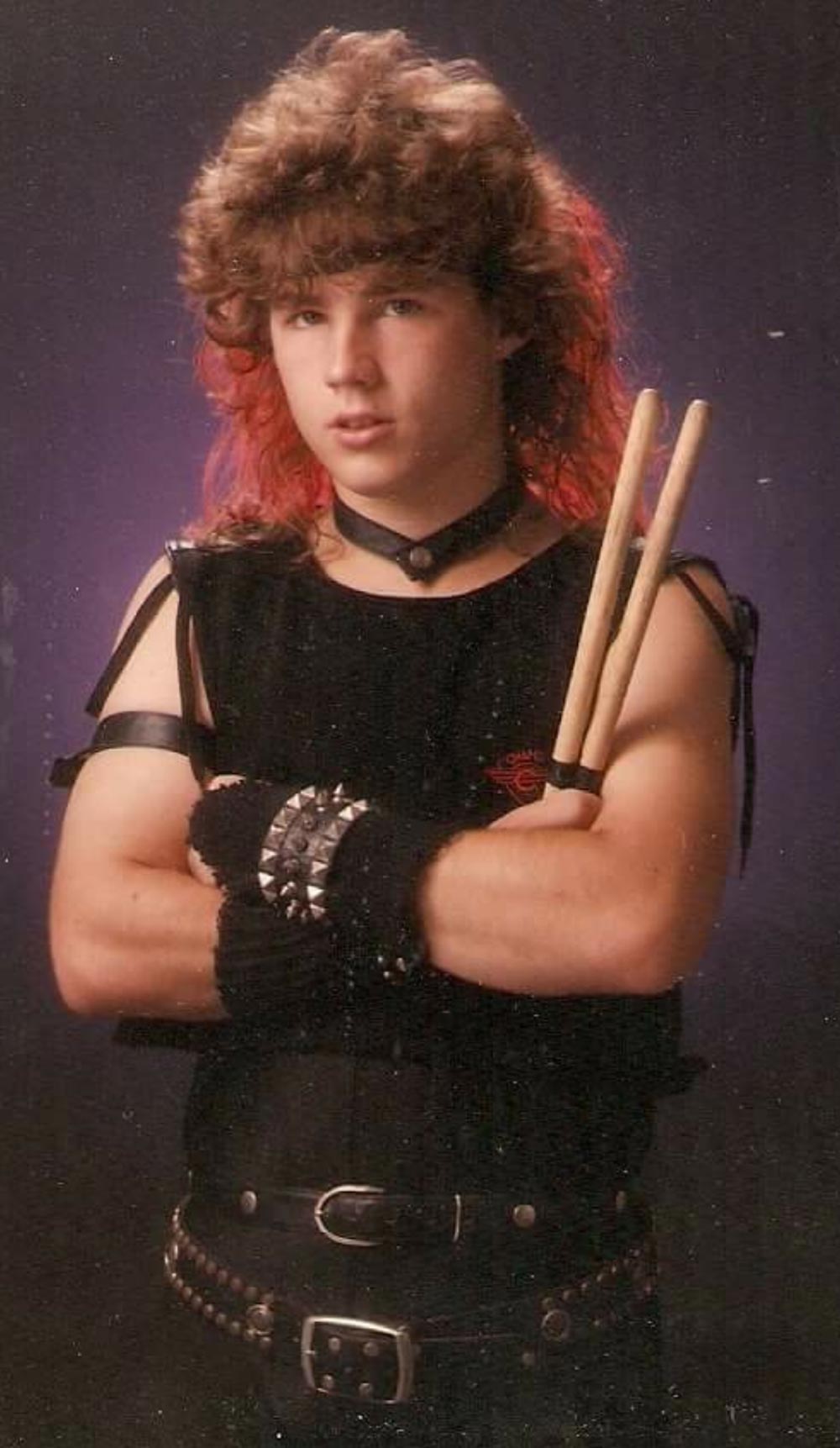 Teenager with a mullet hairstyle, sleeveless black shirt, studded gloves and belt, holding drumsticks, stands with arms crossed against a dark background, evoking an 80s rock or metal style.