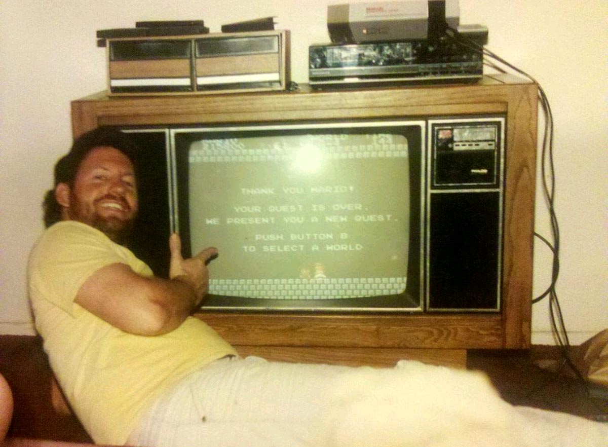 A man with a beard and yellow shirt smiles and points at a retro CRT TV displaying a Thank you Mario! Your quest is over message from a classic video game. An old Nintendo console and other electronics sit on a wooden cabinet above the TV.