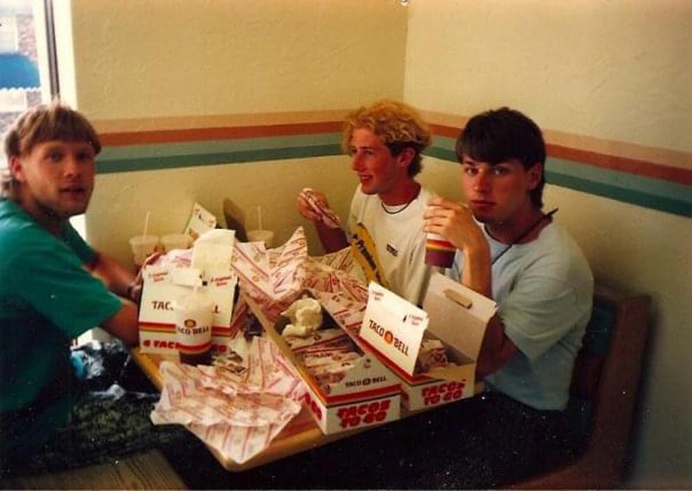 Three young men sit at a Taco Bell booth, surrounded by empty wrappers, drink cups, and food boxes, laughing and eating together. The table is crowded with fast food items.