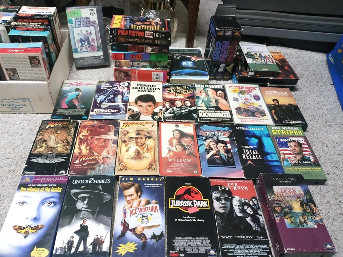 A collection of VHS tapes is spread out on a carpeted floor, featuring popular movies from the 1980s and 1990s, including Jurassic Park, Total Recall, Ace Ventura, Willow, and others, with colorful cover art visible.