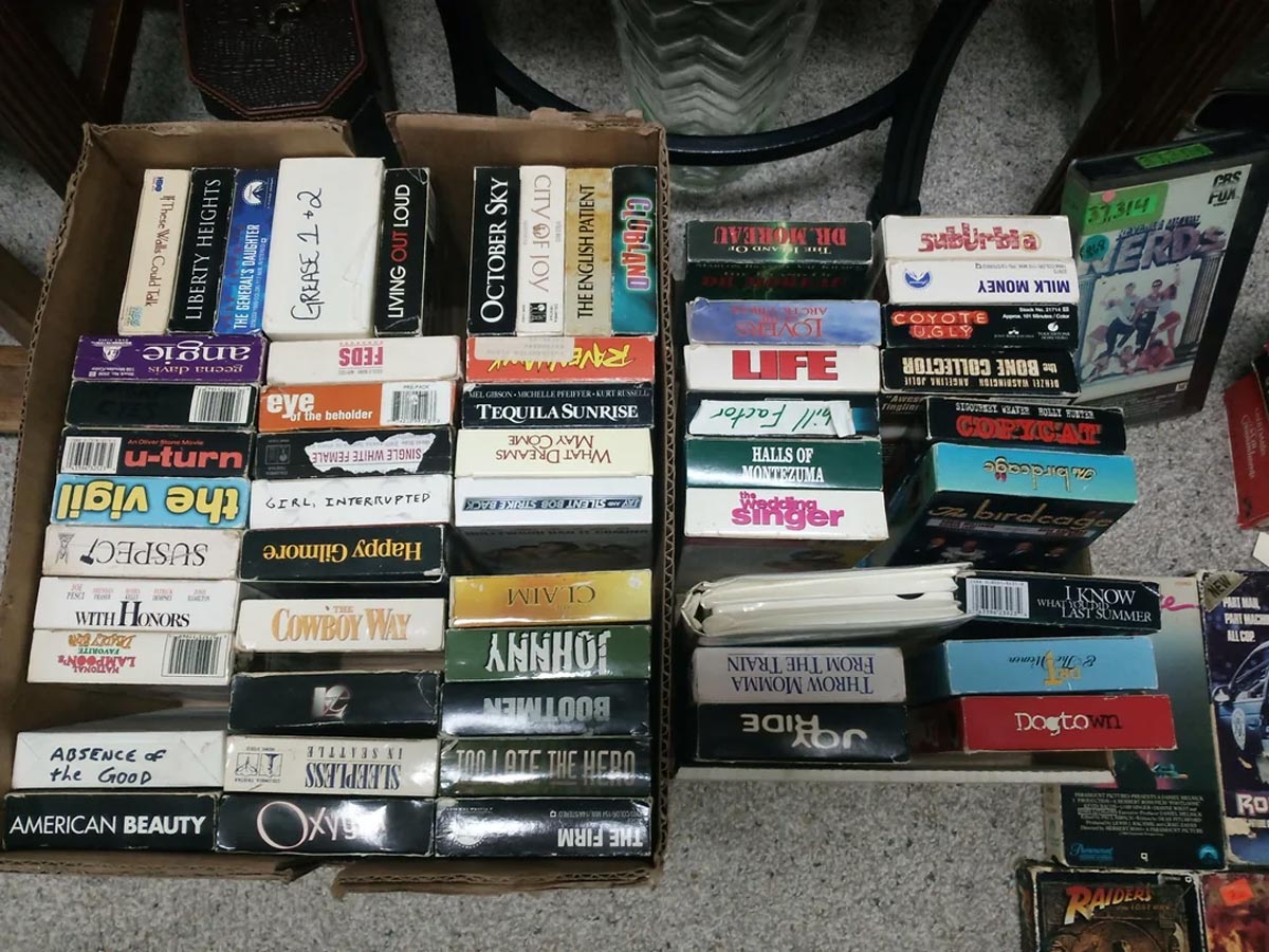 Two cardboard boxes filled with assorted VHS tapes, many stacked vertically and labeled with movie titles such as Grease 2, October Sky, U-Turn, Tequila Sunrise, and American Beauty.