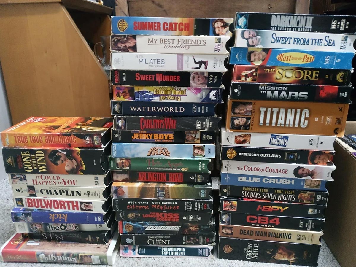 Stacks of VHS tapes arranged in four columns, featuring various movie titles such as Titanic, Waterworld, The Score, Summer Catch, and Chaplin, against a wooden furniture background.