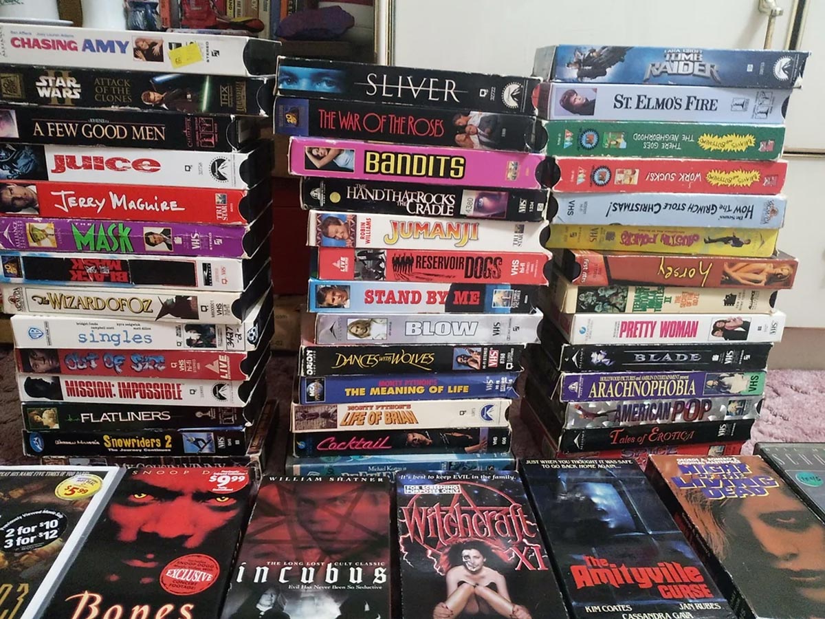 Four stacks of VHS tapes with visible titles, including Star Wars, The Wizard of Oz, Jumanji, Pretty Woman, and Flatliners, sit on a surface with more VHS tapes laid out in front.