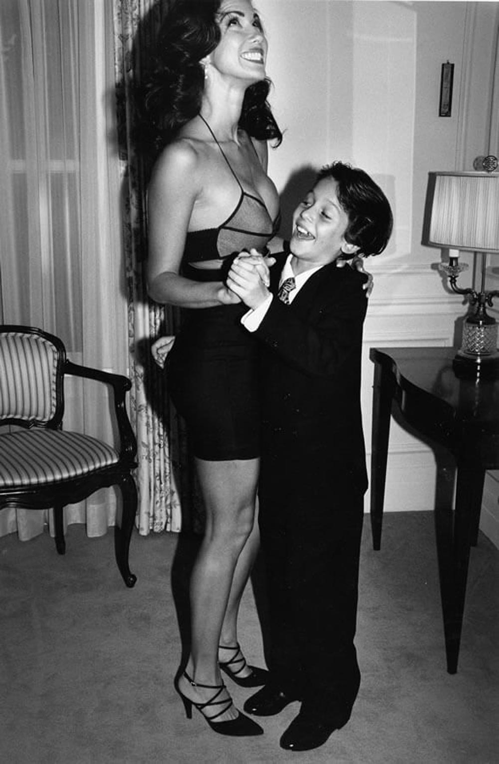 A woman in a black dress and heels dances and smiles with a young boy in a suit and tie in a warmly lit, elegant room with classic furniture.