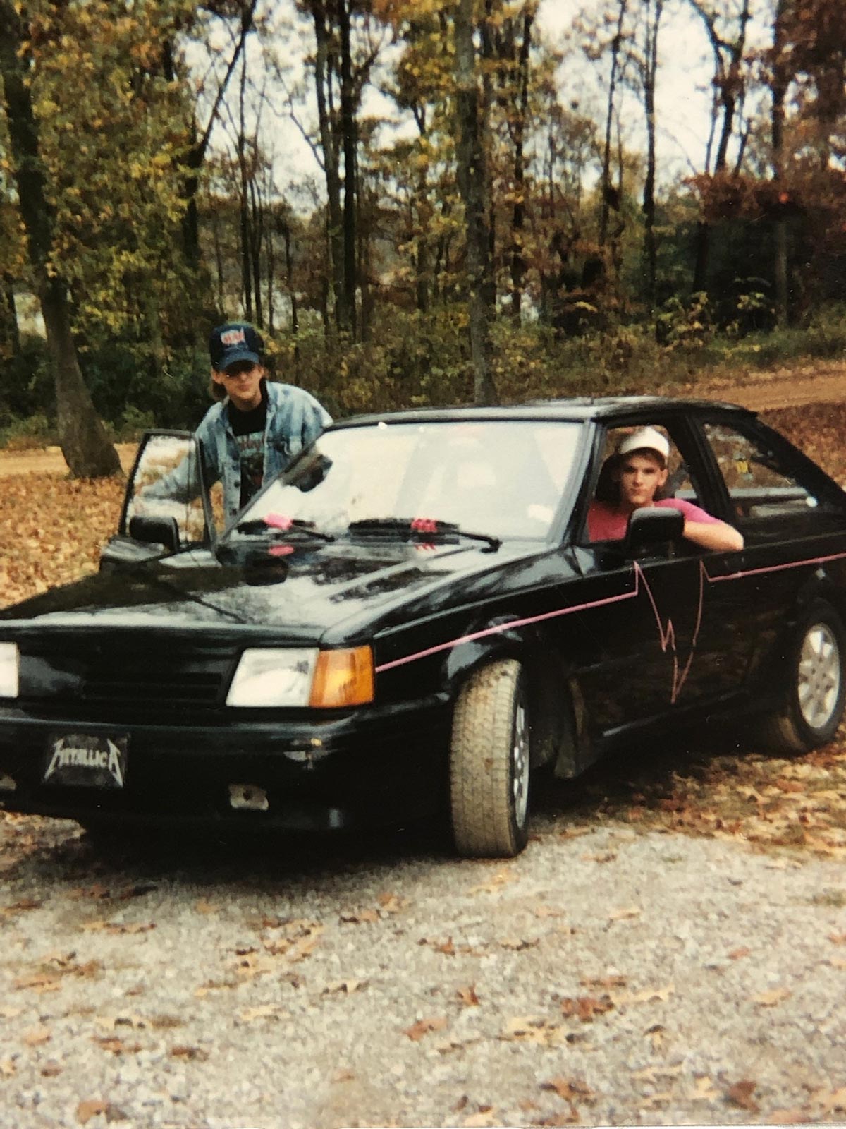 Two young people with hats pose with a black car featuring pink accents and a Metallica license plate. One sits in the drivers seat, while the other leans on the hood. Trees with autumn leaves are in the background.