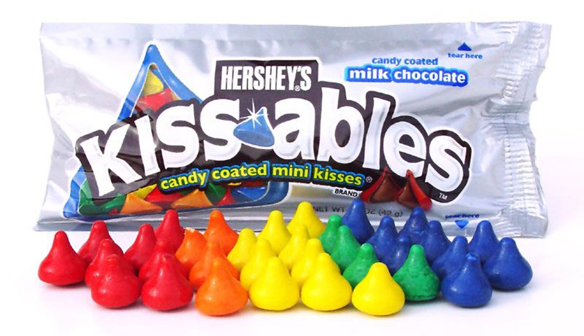A package of Hersheys Kissables candy coated milk chocolate mini kisses with colorful candies—red, orange, yellow, green, and blue—arranged in rows in front of the silver bag.