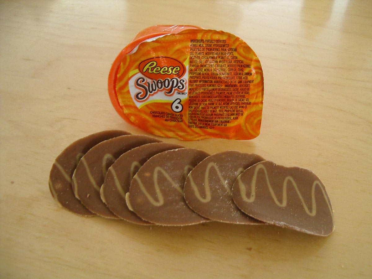 A package of Reeses Swoops with six chocolate pieces arranged in front, each shaped like a potato chip and decorated with a swirl pattern, on a light wooden surface.