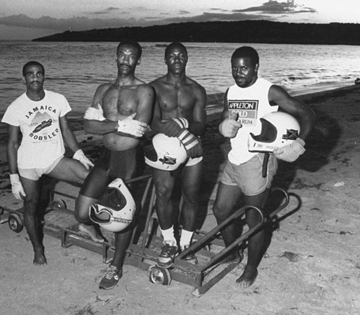 Four men stand on a beach at sunset beside a bobsled, holding helmets. Three wear athletic gear with Jamaica Bobsled visible on one shirt, referencing the Jamaican bobsled team. A body of water and hills are in the background.