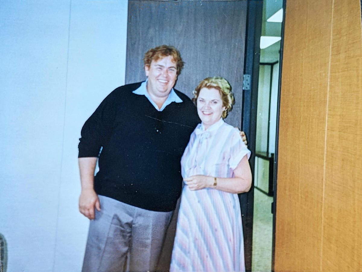 A man in a black sweater and gray pants stands next to a woman in a striped dress. They are smiling and posing for the camera in an office hallway near a wooden door.