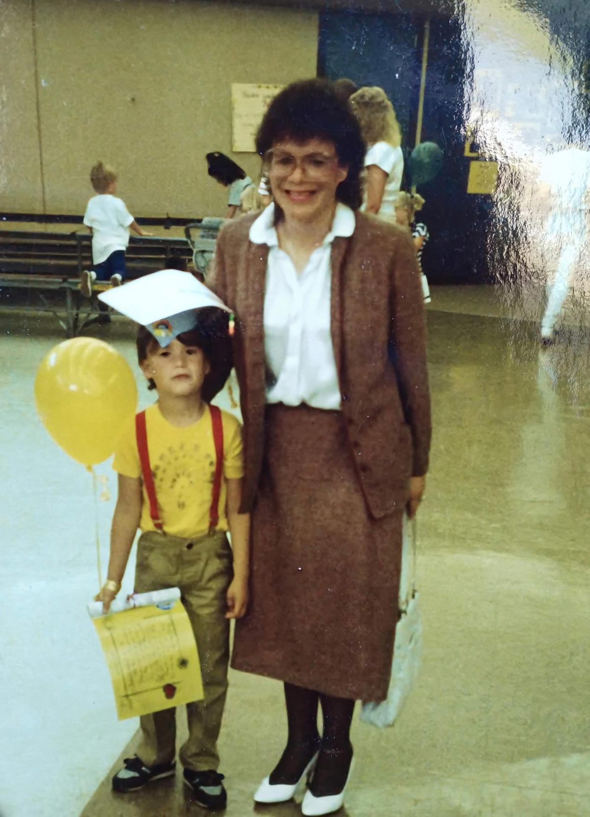 A young boy in a graduation cap, yellow shirt, and red suspenders holds yellow balloons and a certificate while standing next to a smiling woman in a pink suit and glasses indoors.