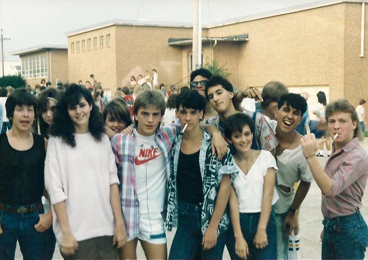 A group of teenagers, some smiling and some with serious expressions, pose together outside a school building. A few hold cigarettes. Other students and buildings are visible in the background.