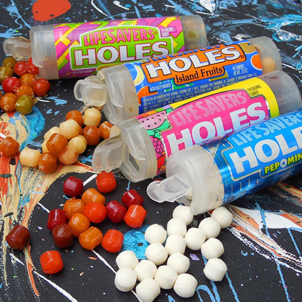 Four containers of Life Savers Holes candy, each with a different flavor, are open and spilling colorful, round candies onto a multicolored surface. The candies vary in color and flavor.