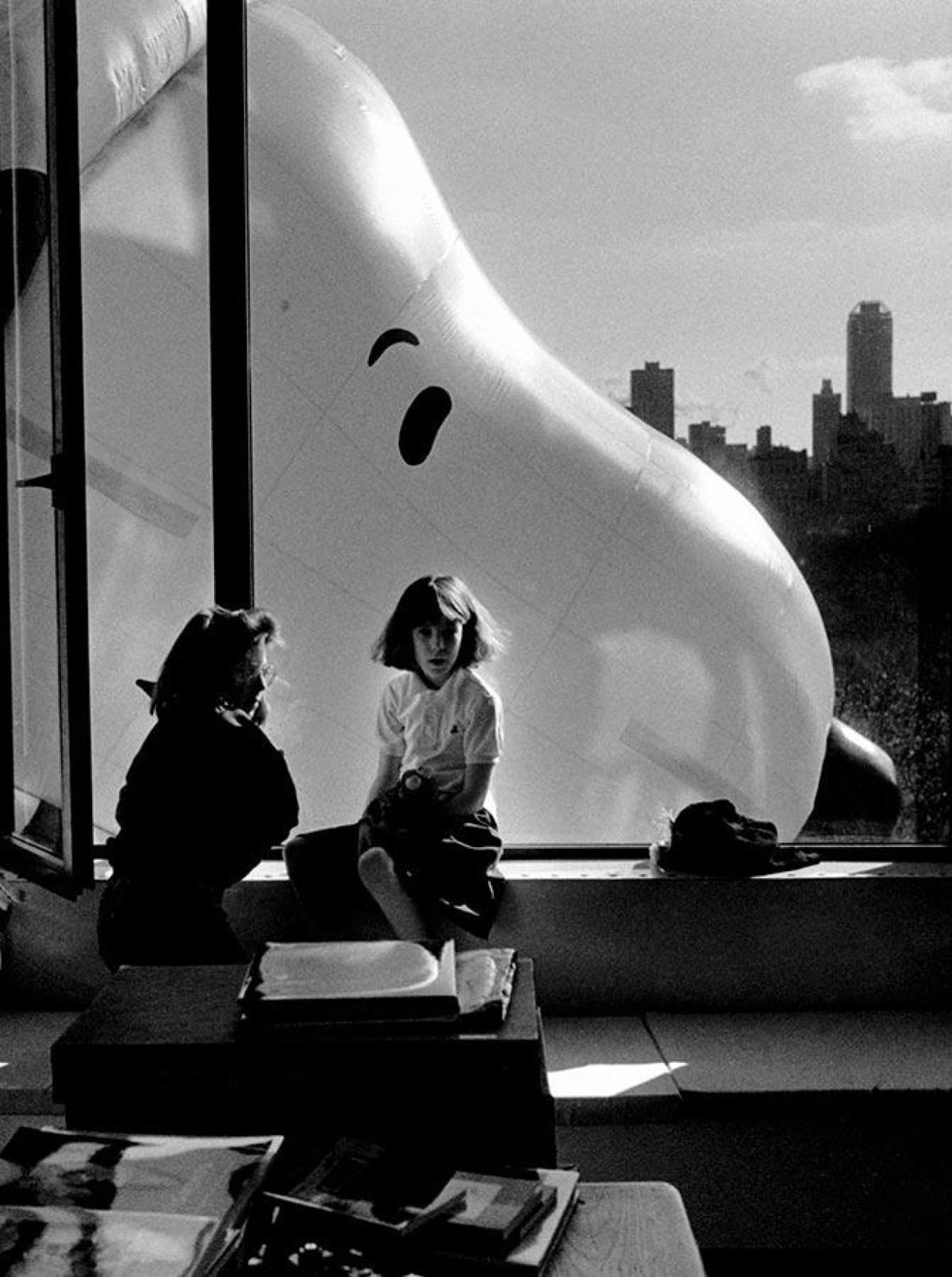 Two people sit by a large window as a giant Snoopy balloon floats outside, filling most of the view. City buildings and trees are visible in the background, and sunlight streams into the room.