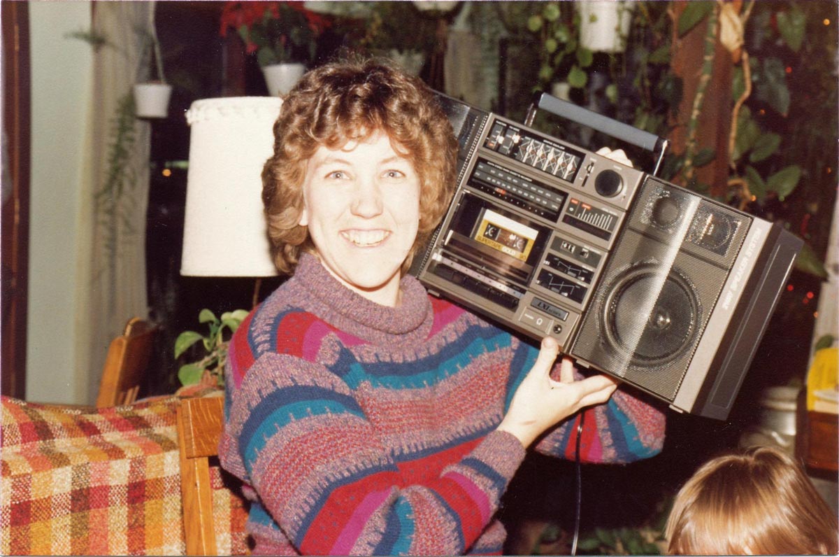 A smiling woman in a colorful striped sweater holds a large vintage boombox on her shoulder, sitting indoors on a patterned chair with plants in the background.