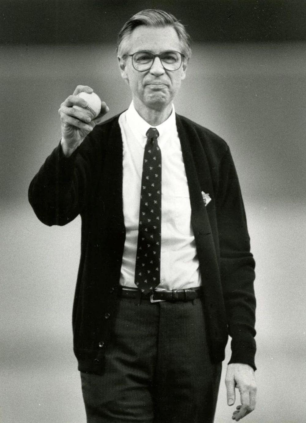An older man wearing glasses, a tie, and a dark cardigan holds up a baseball in one hand, standing and facing the camera with a slight smile in a black-and-white photo.