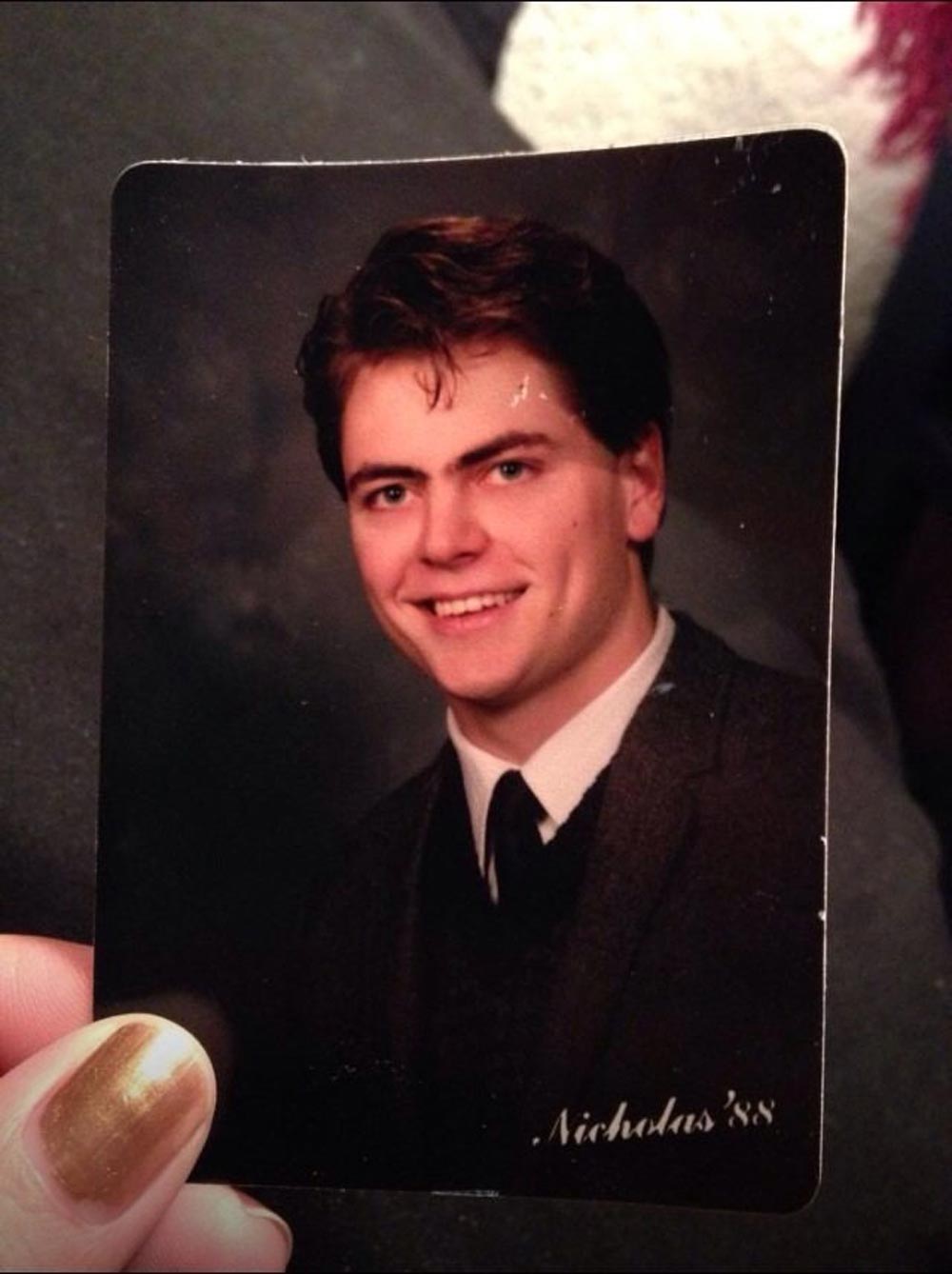 A hand holding a portrait photo of a young man in a suit and tie, smiling at the camera. The name “Nicholas ’88” is written in cursive in the lower right corner of the photograph.