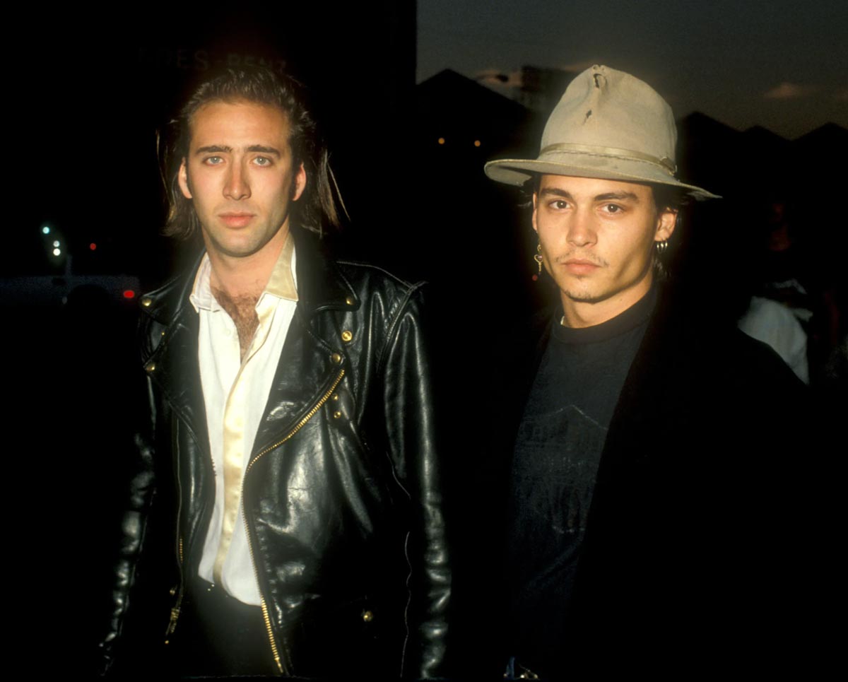 Two men stand side by side at night. One wears a black leather jacket over a white shirt, and the other wears a light-colored fedora hat, a dark coat, and a dark shirt. Both look directly at the camera.