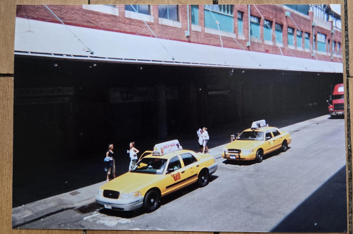 Two yellow taxis are parked on a city street near a sidewalk where five people stand. The background features a brick building with a long awning, and a red truck is visible further down the street.