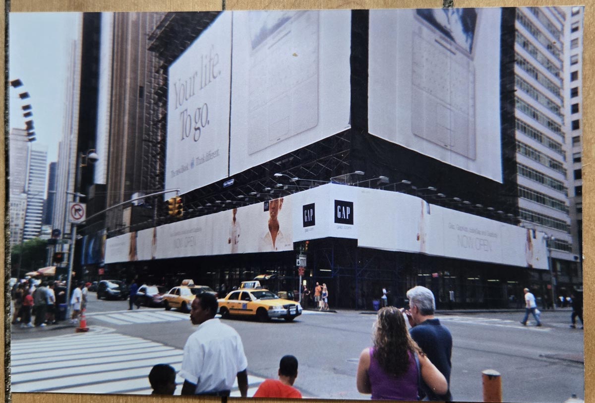 People cross a busy New York City street near yellow taxis. The corner building is wrapped in large GAP advertisements, with a sign reading “You like yoga.” Skyscrapers rise in the background under a clear sky.
