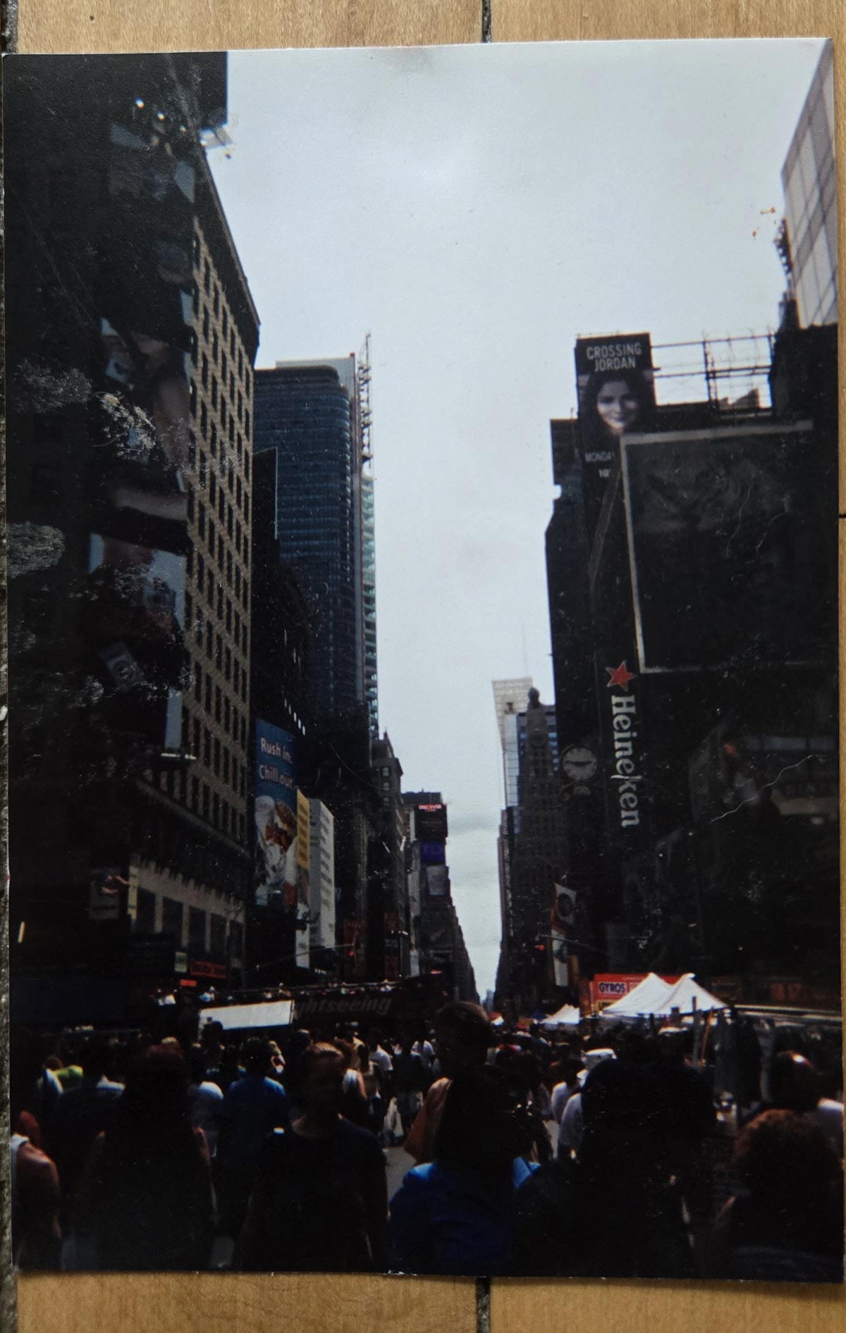 A large crowd walks down a busy street lined with tall buildings and billboards in an urban area, likely Times Square in New York City, on a sunny day.