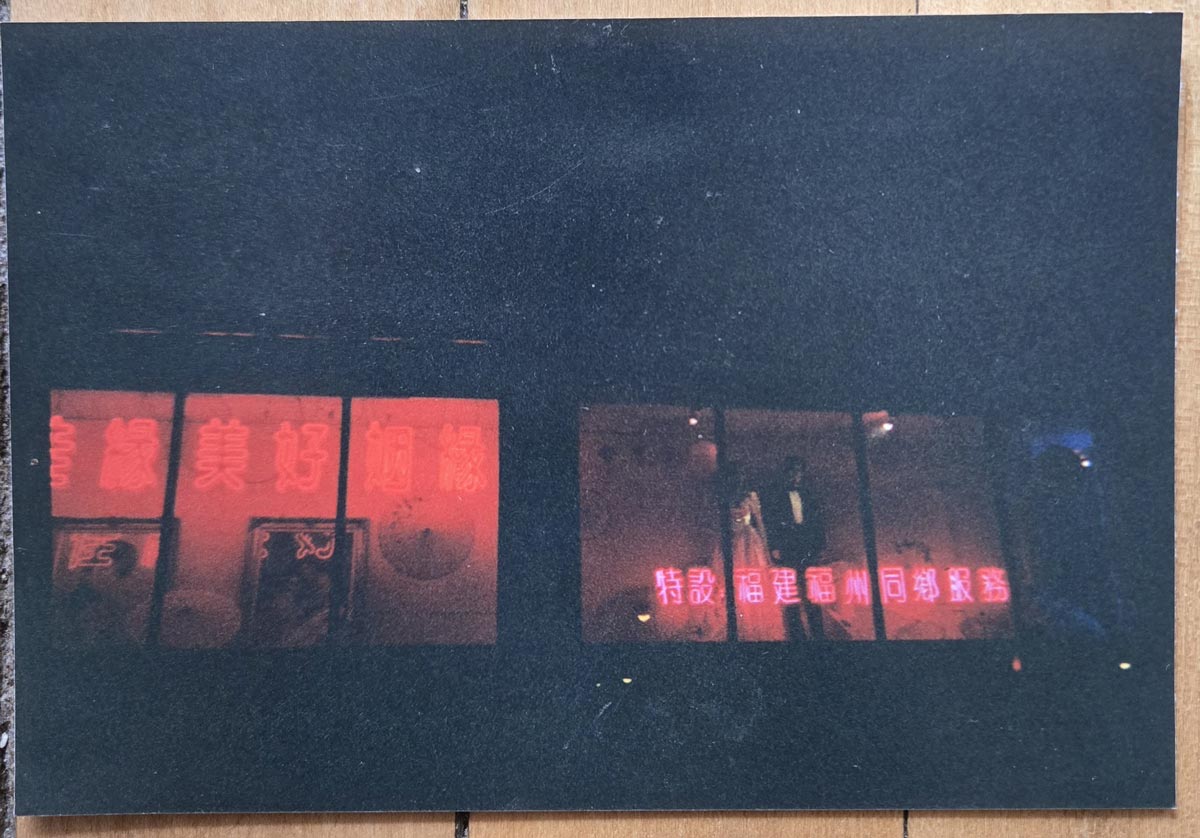 A dark photo showing bright red neon Chinese characters glowing in shop windows at night, with some posters visible below the signs. The rest of the scene is obscured by darkness.