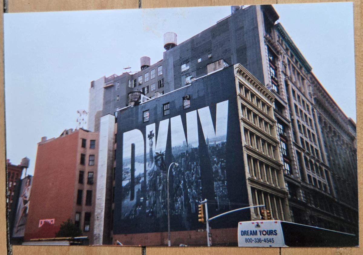 A large DKNY billboard covers the side of a city building, with a black-and-white cityscape in the background. Other tall buildings surround it, and a Dream Tours sign is visible in the bottom right corner.