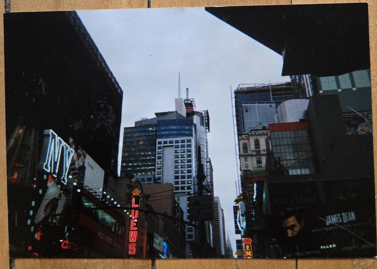 A cityscape view of Times Square in New York City, featuring tall buildings, bright neon signs, billboards, and a cloudy sky overhead.
