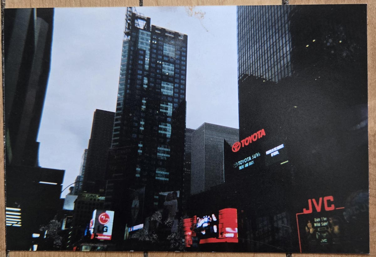 A cityscape featuring tall skyscrapers and electronic billboards, including prominent Toyota and JVC signs, on a cloudy day. The scene has an urban, bustling atmosphere.