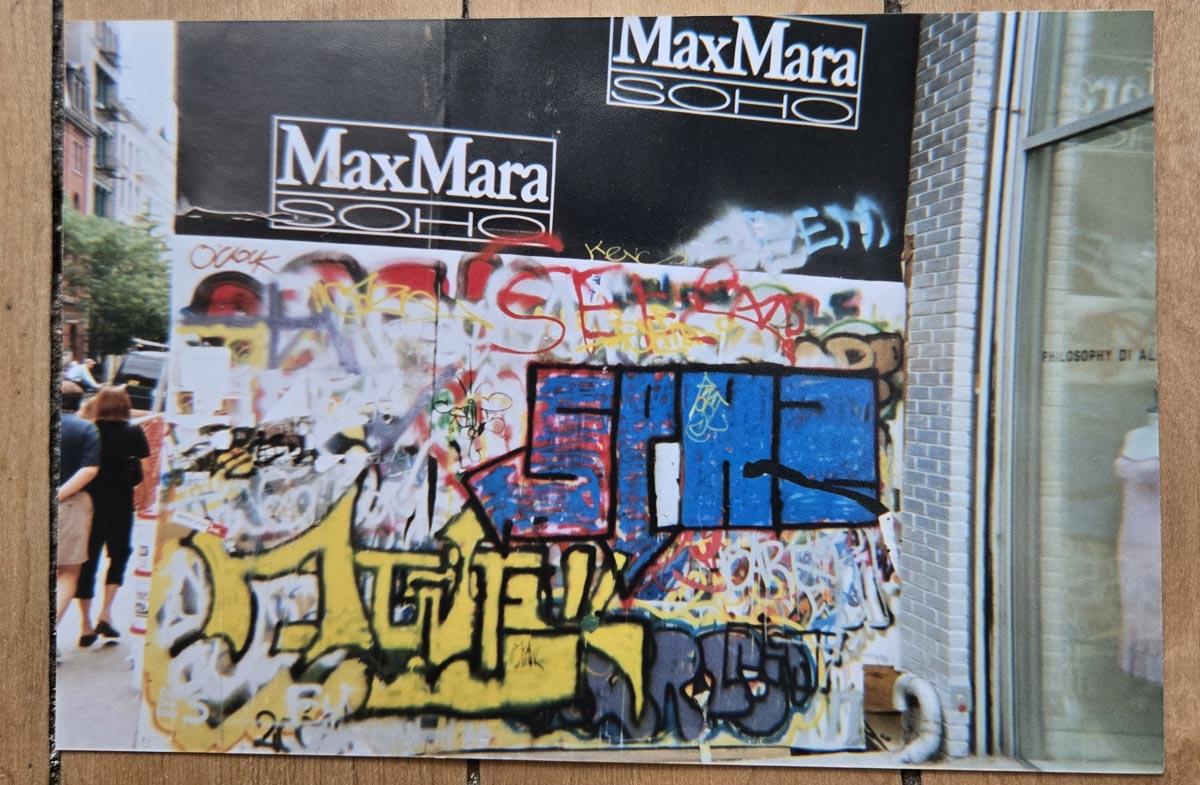 A wall covered in colorful graffiti and tags below two MaxMara SOHO signs; people walk on the sidewalk and a store window reflects the street scene.