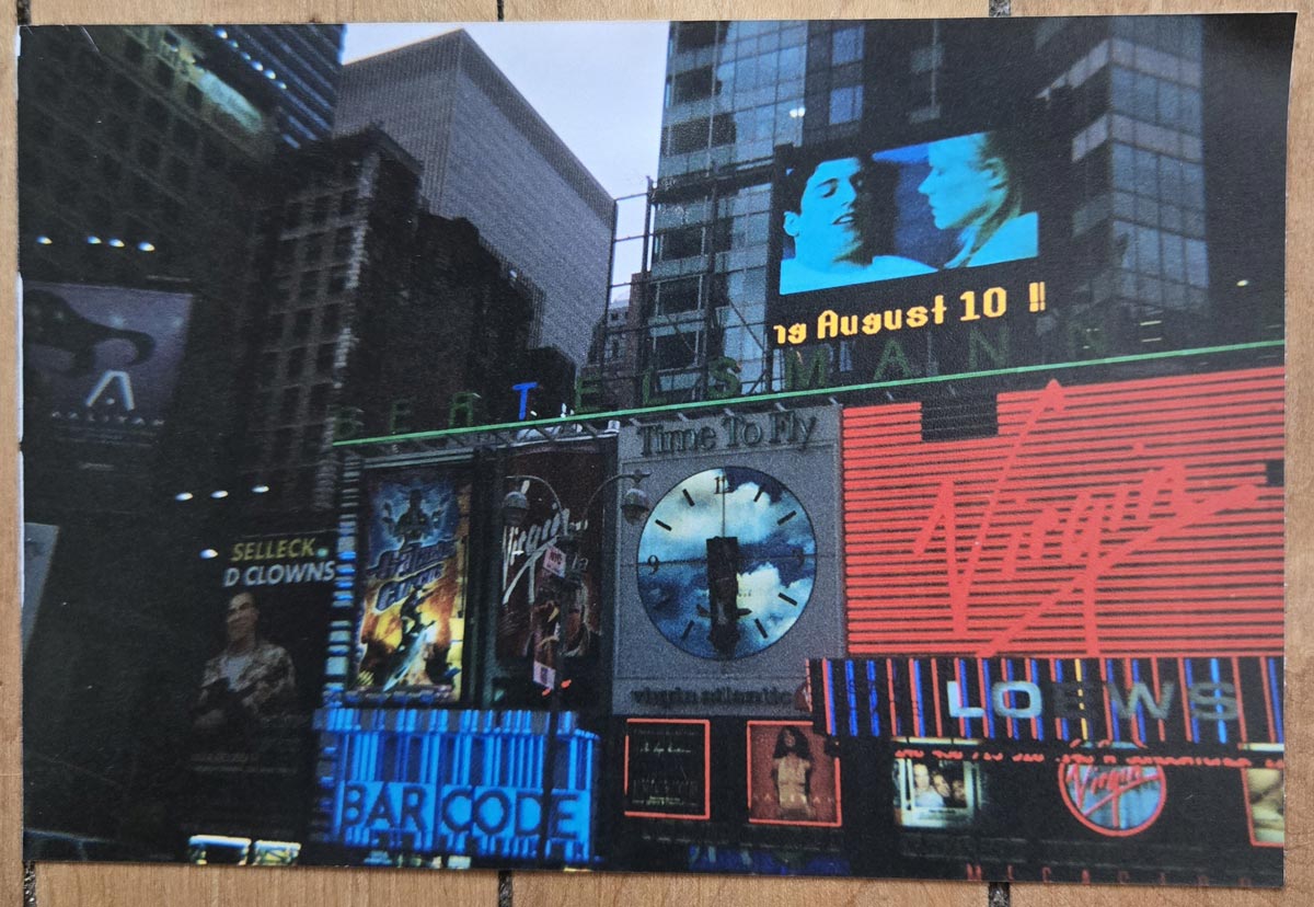 Times Square scene with bright neon signs, including a large Virgin logo, a Bar Code sign, a clock, movie posters, and a video screen displaying two people and the text 19 August 10!! against tall city buildings.