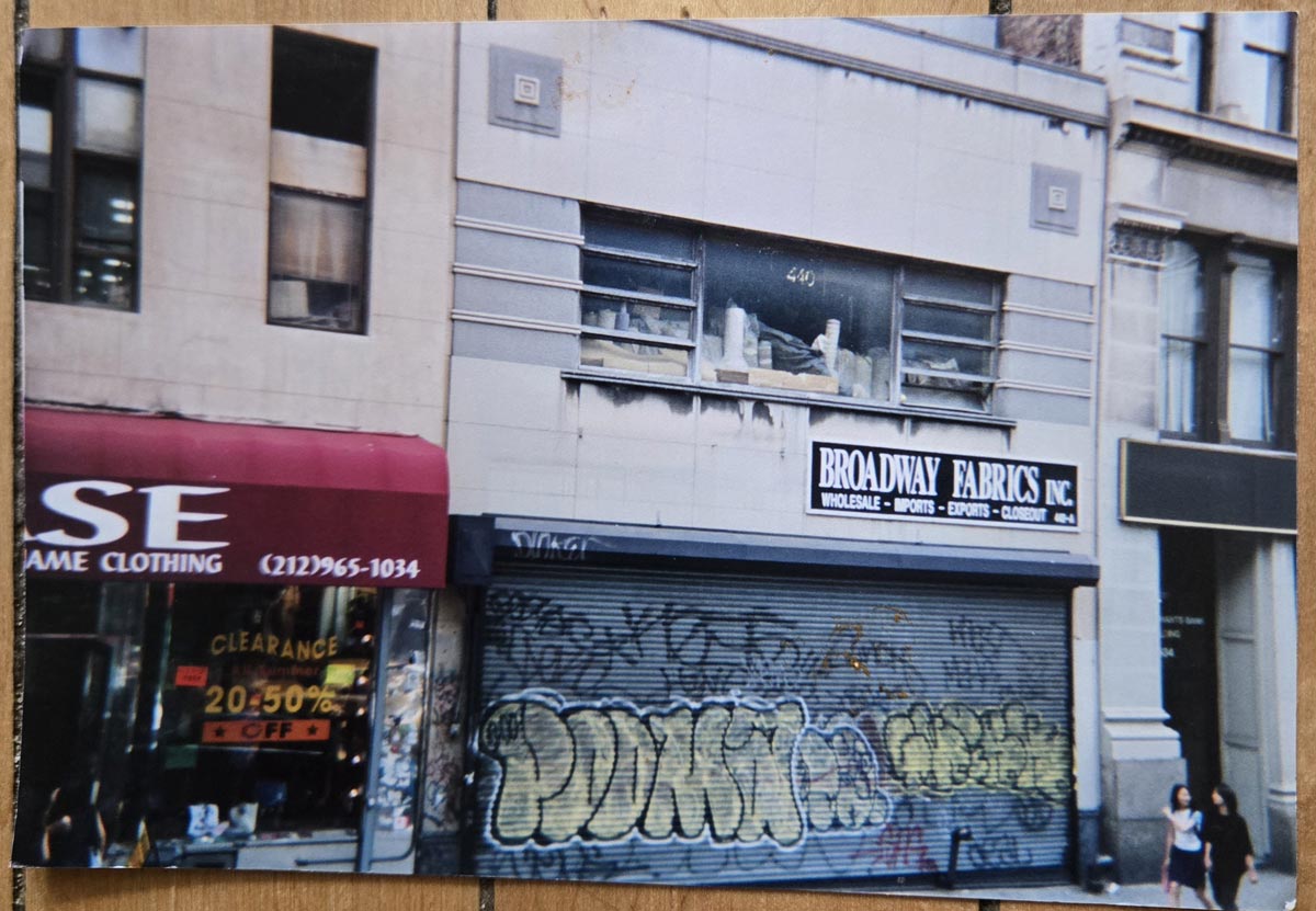 A shuttered storefront with graffiti covers the entrance to Broadway Fabrics, Inc. Next door, a clothing store displays a red awning and a Clearance 20-50% off sign in the window.