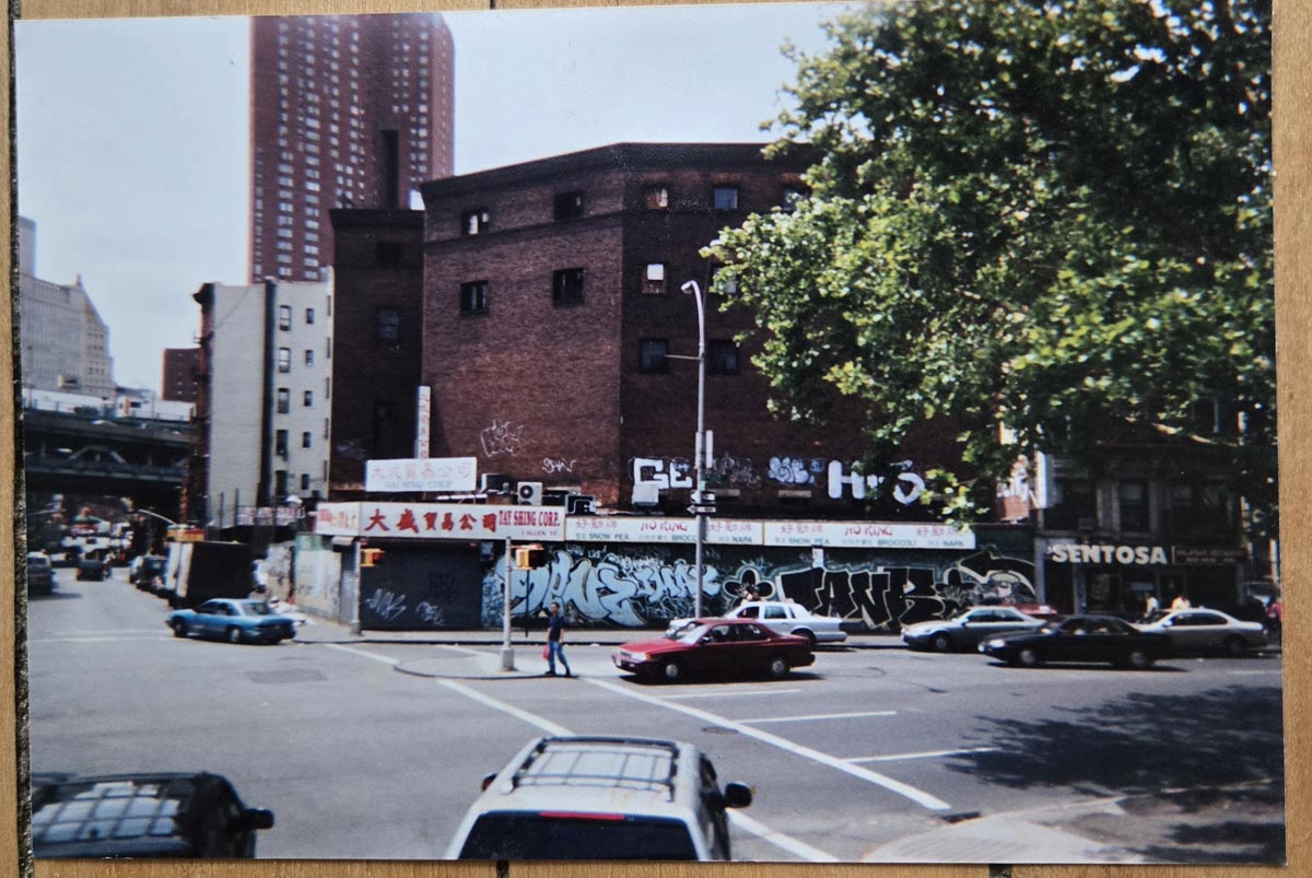 A city street corner with cars driving by, pedestrians crossing, and brick buildings covered in graffiti. Storefronts with Asian signage are visible, along with a tree and a tall red building in the background.