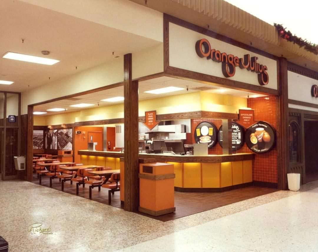 A vintage Orange Julius store in a mall with orange and yellow décor, empty orange chairs and tables, and a counter with menu signs on the wall behind it.