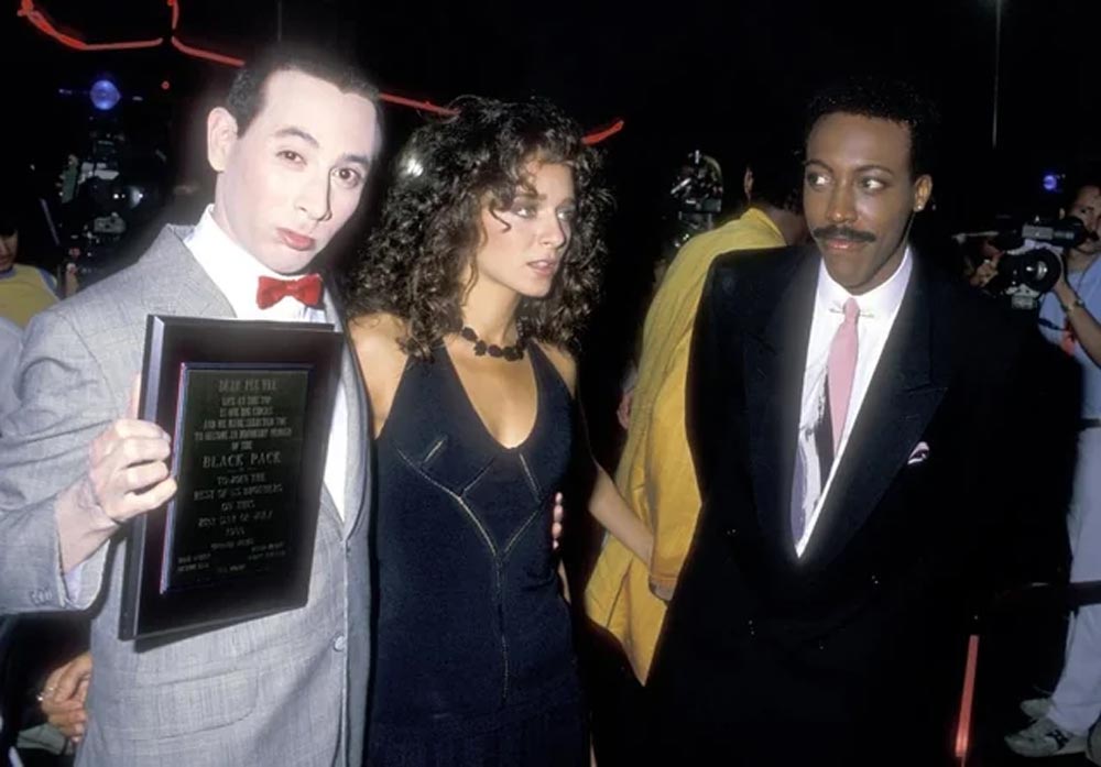 Three people stand together at an event; one holds a plaque, another wears a dark dress with curly hair, and the third wears a suit and tie. There are cameras and people visible in the background.
