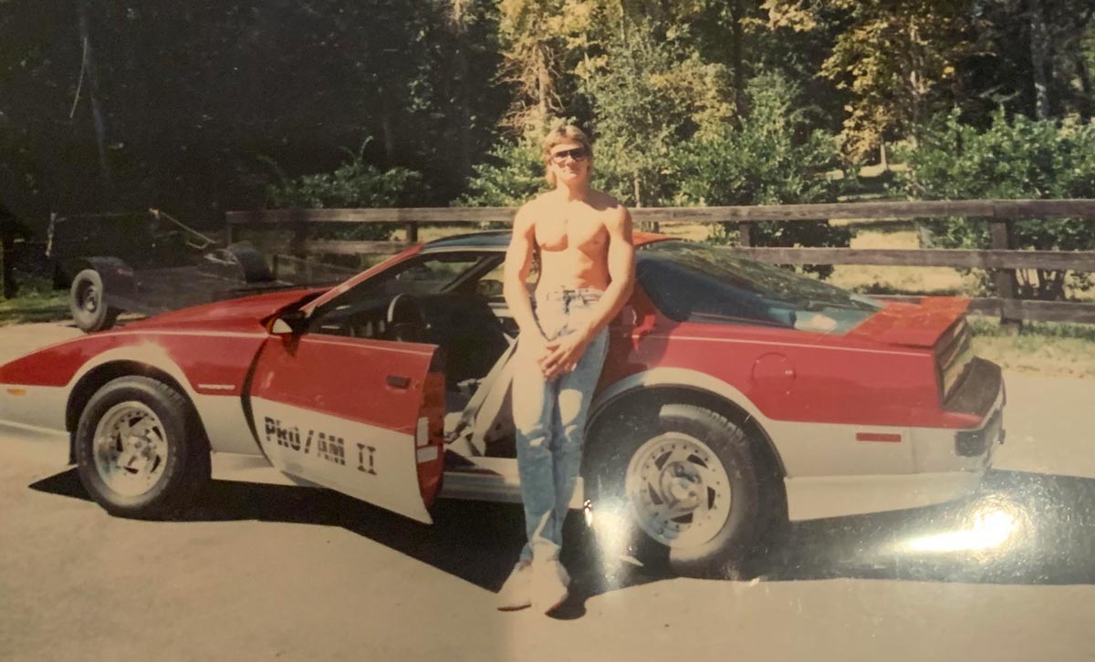 A shirtless man wearing sunglasses and jeans stands in front of a red and white sports car with an open door, outdoors on a sunny day with trees and a wooden fence in the background.