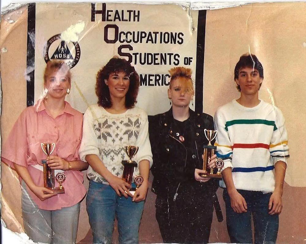 Four young people stand in front of a Health Occupations Students of America sign, holding trophies. Three are smiling; one with a mohawk and leather jacket has a neutral expression. The photo appears old and worn.