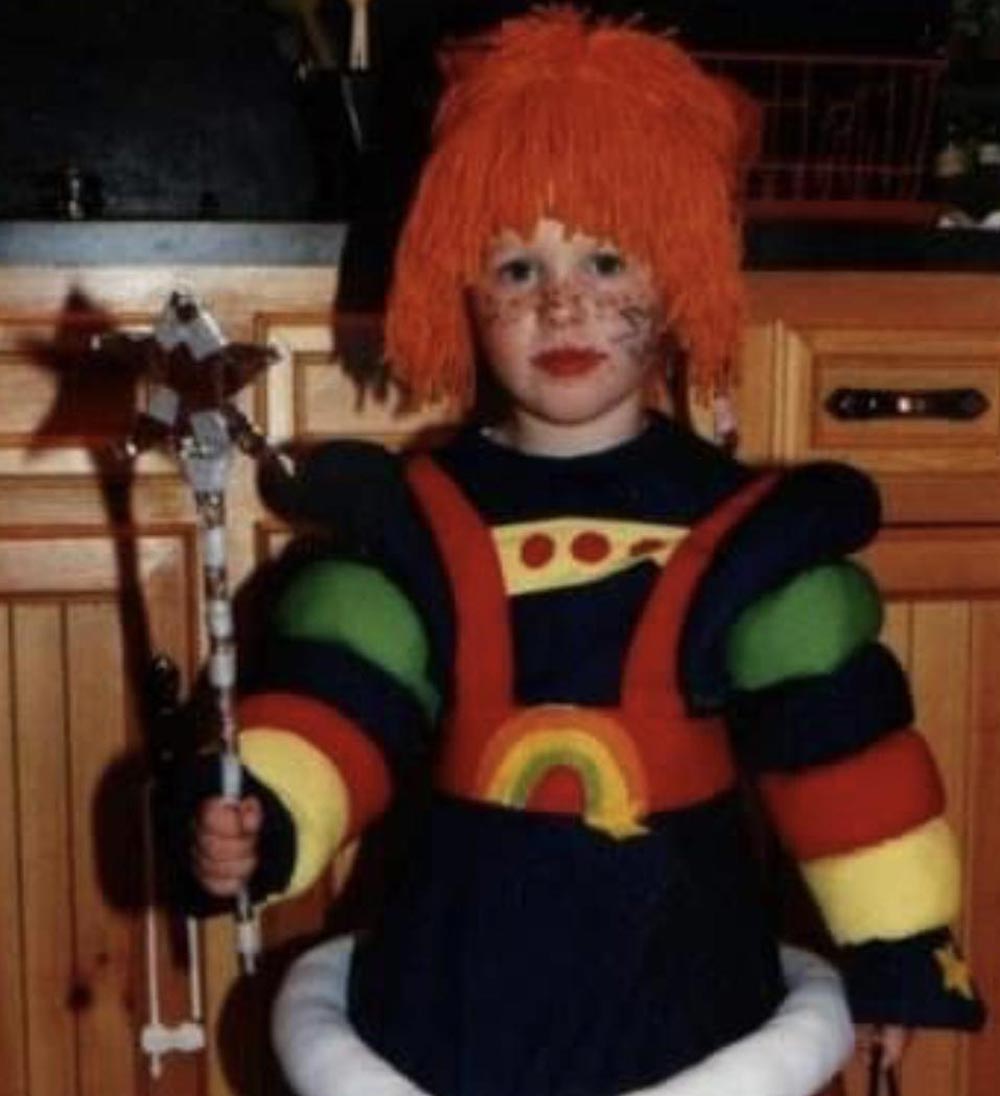 A young child in a colorful, padded costume with rainbow-striped sleeves, a red yarn wig, face paint, and holding a staff, standing indoors in front of wooden cabinets.