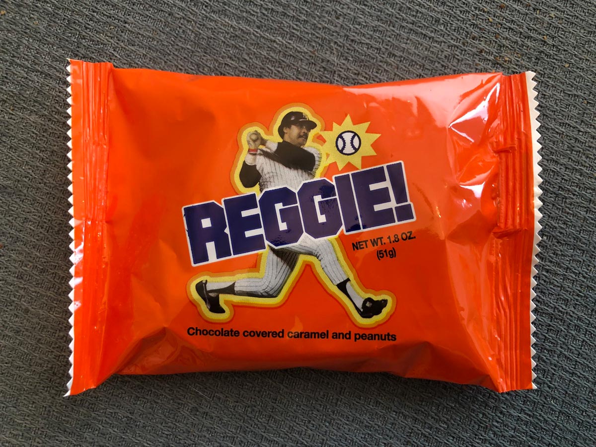 An orange wrapper labeled REGGIE! with an illustration of a baseball player swinging a bat. The text states Chocolate covered caramel and peanuts and lists the net weight as 1.8 oz (51g).