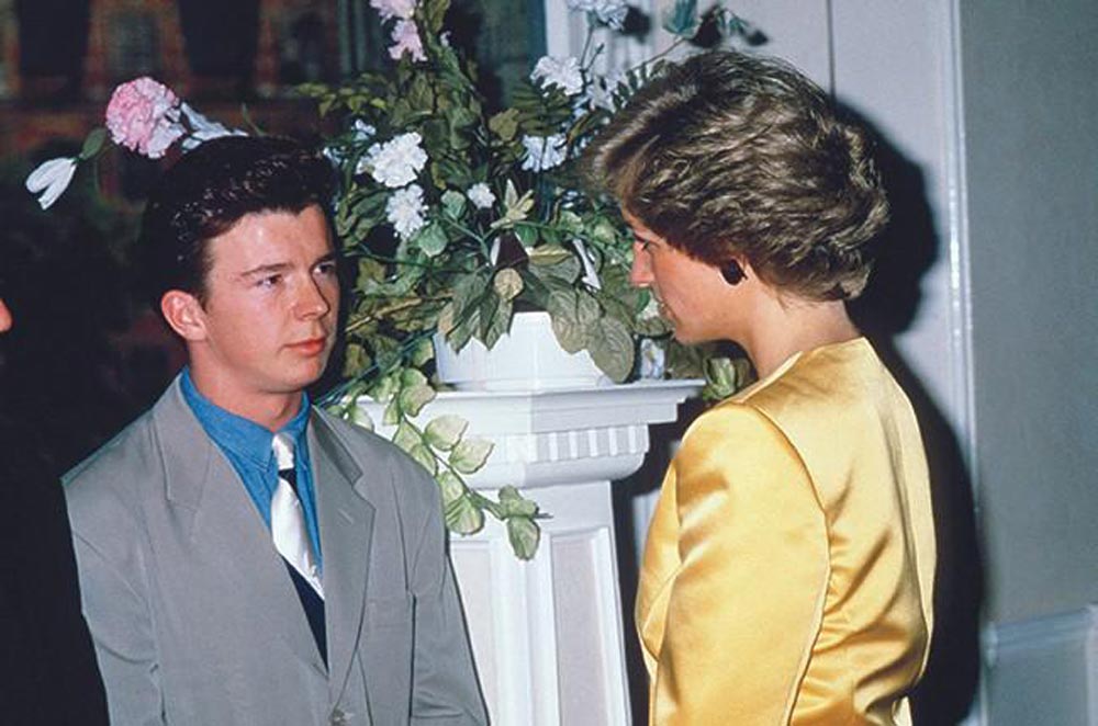 A young man in a gray suit and blue tie stands next to a woman in a yellow dress jacket. They are indoors, conversing near a white pillar decorated with pink and white flowers.