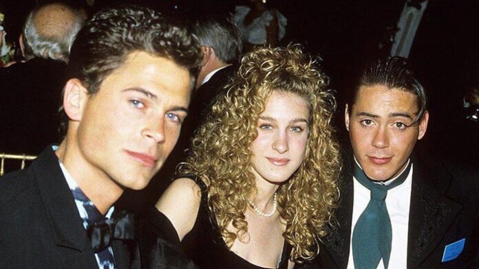 Three young adults dressed formally sit closely together at an event, looking at the camera. The background is dimly lit with other people visible behind them.